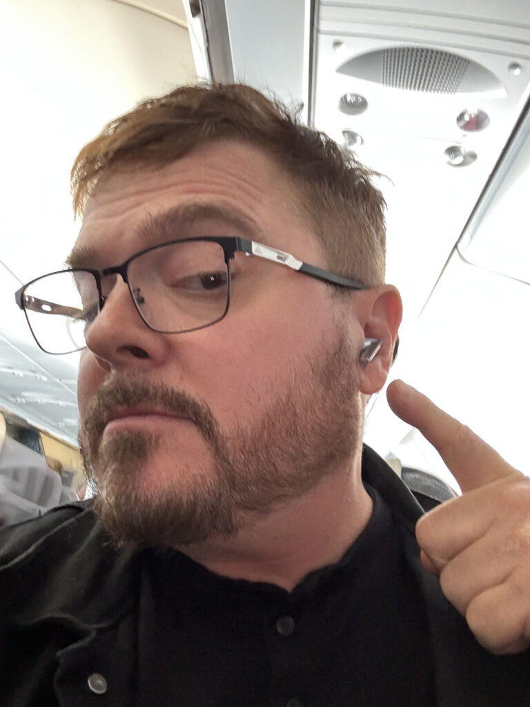 Man wearing glasses pointing to his ear, which has a device in it.