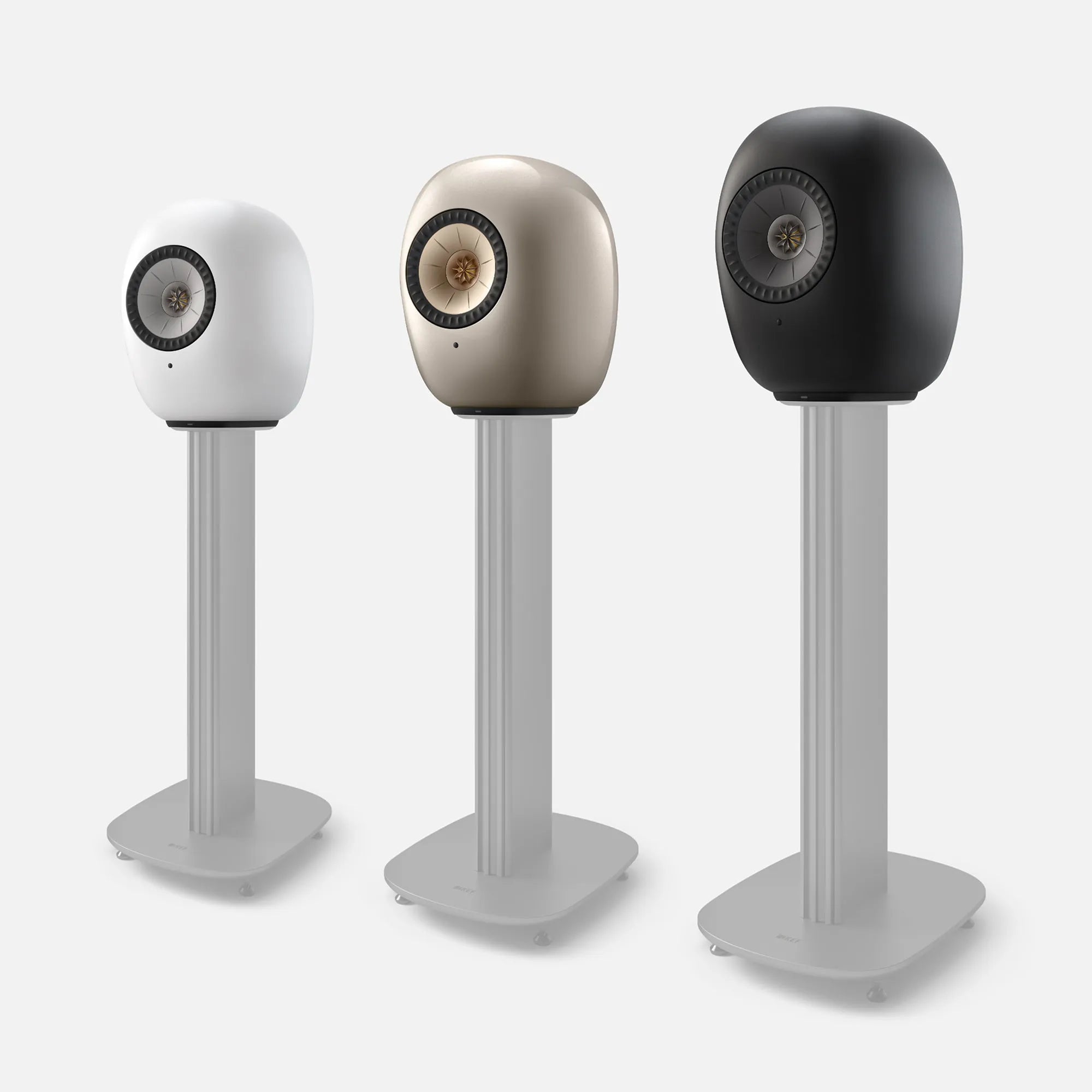 Three KEF LS LUXE wireless speakers in white, titanium, and black on matching floor stands.