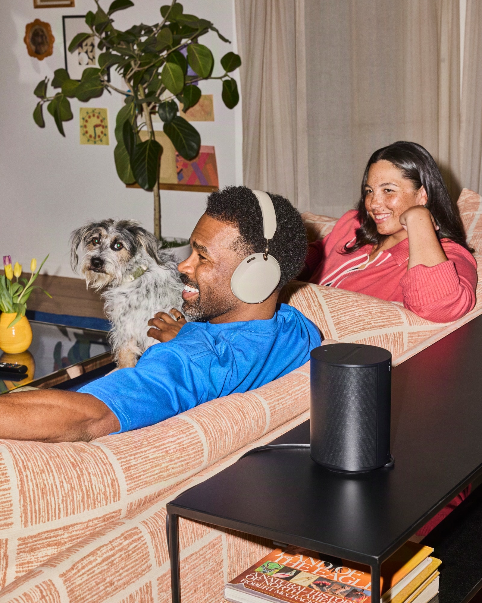 A person wearing white Sonos Ace Ultra headphones beside a Sonos speaker in a living room.