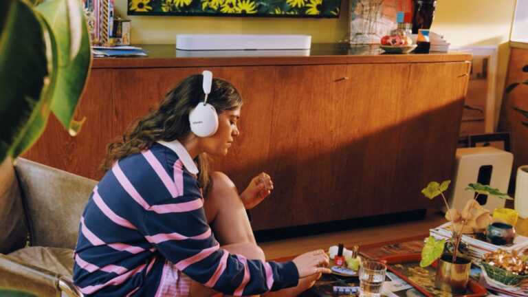 A person wearing white Sonos Ace Ultra headphones with a white Beam Ultra soundbar in the background.
