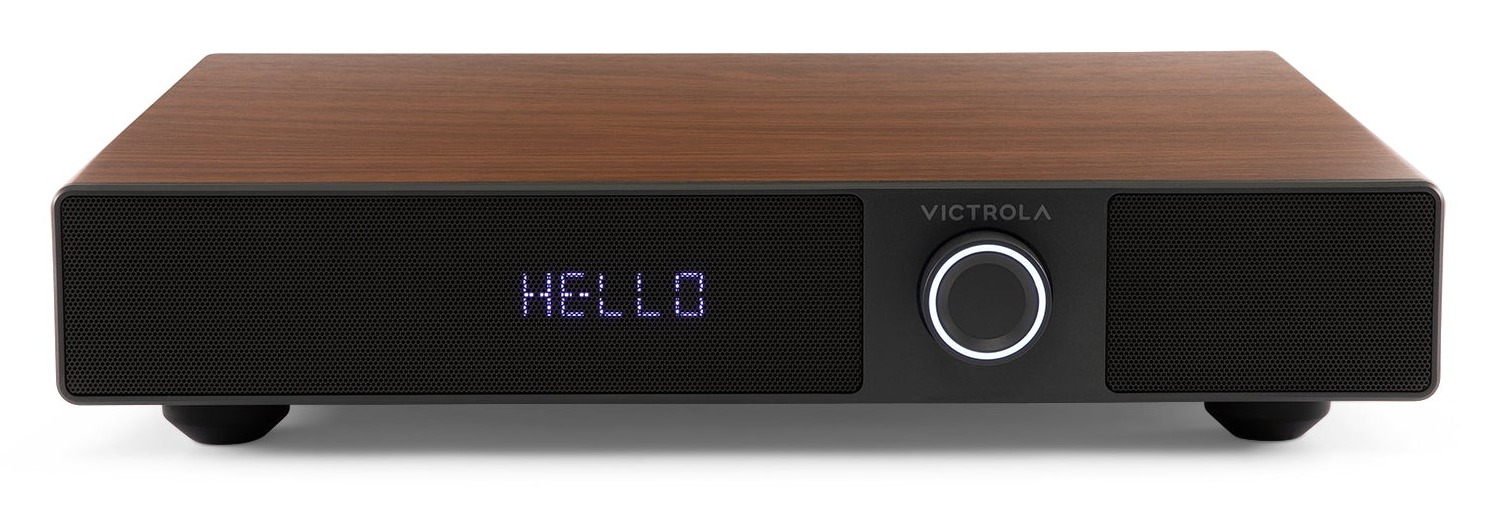 Front view of the Walnut Victrola Soundstage speaker base showing the illuminated HELLO LED display behind the mesh grille and the central rotary control knob